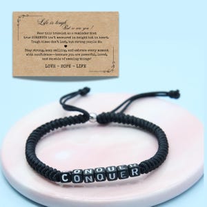 Op de afbeelding: Zwart macrame armband met het woord "CONQUER" gespeld in witte letterkralen. Op de achtergrond een kleine kaart met inspirerende tekst. Op de kaart staat "Het leven is zwaar, maar jij ook!"