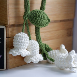 May include: A crocheted lily of the valley flower garland with white flowers and green leaves. The garland is hanging from a wooden box with the text "Mees" on the front.