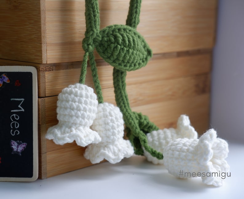 May include: A crocheted lily of the valley flower garland with white flowers and green leaves. The garland is hanging from a wooden box with the text "Mees" on the front.