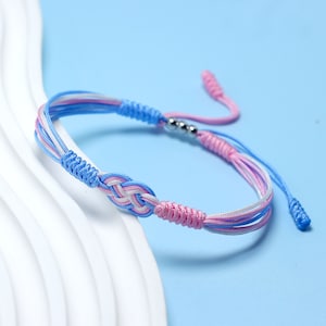 Puede incluir: Un brazalete de amistad trenzado azul, rosa y blanco con un acento de cuentas plateadas. El brazalete está atado con un nudo y tiene una borla larga.
