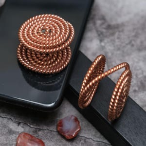 May include: Two copper-colored phone grips featuring a circular, coiled rope-like design. One grip is attached to the back of a black smartphone, while the other stands upright, showcasing its cylindrical, spiraled structure and metallic finish.