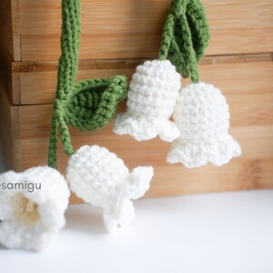 May include: A string of crocheted white lily of the valley flowers with green leaves. The flowers are hanging from a wooden surface.