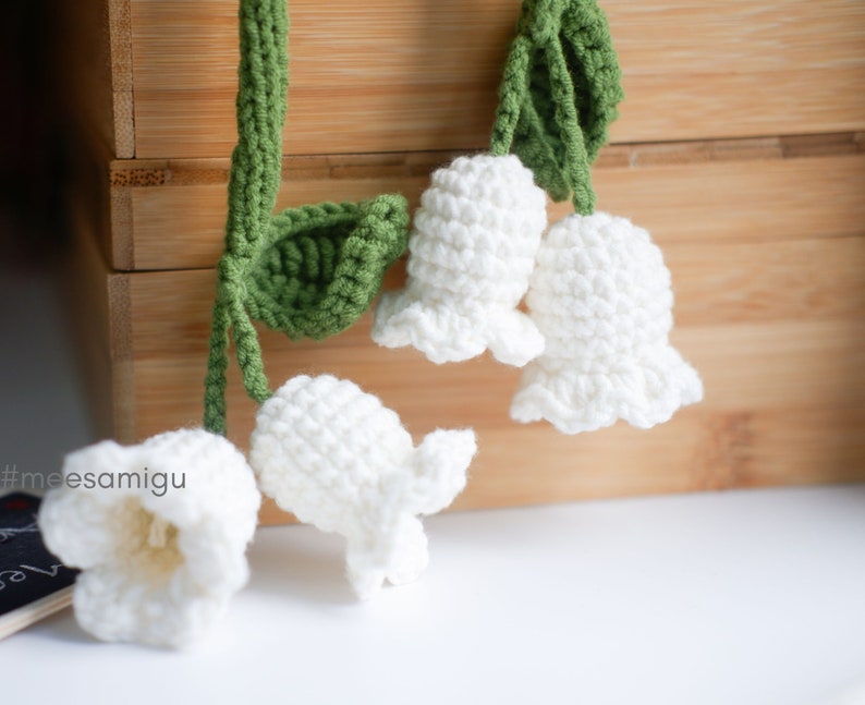 May include: A string of crocheted white lily of the valley flowers with green leaves. The flowers are hanging from a wooden surface.