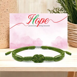 Puede incluir: Pulsera de cuerda verde con un nudo en el centro. La pulsera tiene una cuenta plateada a cada lado del nudo. La pulsera está sobre una superficie de madera. El texto "Hope protection string bracelet" está sobre un fondo rosa detrás de la pulsera.