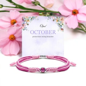 Puede incluir: Pulsera de protección de cuerda rosa con una piedra preciosa de ópalo y detalles plateados. La pulsera se muestra en una tarjeta con el texto "Opal OCTOBER protection string bracelet".