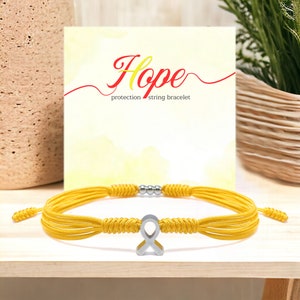 Peut inclure: Un bracelet de corde jaune avec un charm en forme de ruban argenté. Le bracelet est présenté devant une carte avec le mot "Hope" et le texte "protection string bracelet". Le bracelet est sur une surface en bois.