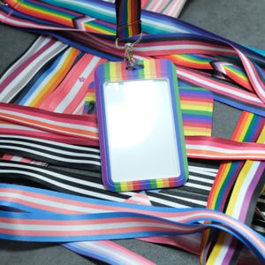 May include: A rainbow lanyard with a blank ID badge holder. The lanyard is made of multiple colorful stripes, including red, orange, yellow, green, blue, indigo, and violet. The ID badge holder is clear plastic with a rainbow border.