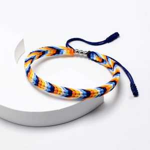 Könnte beinhalten: Ein farbenfrohes geflochtenes Armband mit blauen, gelben, orangen und weißen Fäden. Das Armband hat eine silberne Metallperle und eine blaue Schnur mit einem Knoten am Ende.