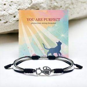 Puede incluir: Pulsera de cuerda negra y blanca con un dije de gato plateado. La pulsera está sobre un fondo blanco con una tarjeta que dice "You are Purrfect protection string bracelet".