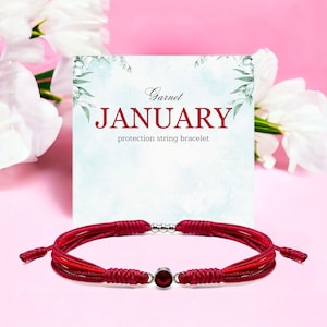 May include: Red string bracelet with a garnet gemstone and silver beads. The bracelet is tied with a knot and has a small silver charm. The bracelet is on a pink background with a card that says "Garnet JANUARY protection string bracelet".