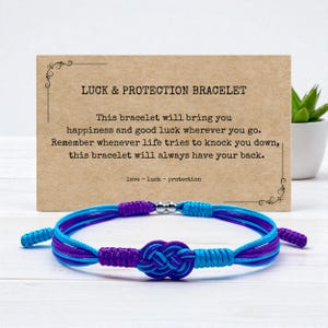 May include: A blue and purple woven bracelet with a silver bead clasp. The bracelet is displayed in front of a card that reads "LUCK & PROTECTION BRACELET". The card also includes text about happiness and good luck.