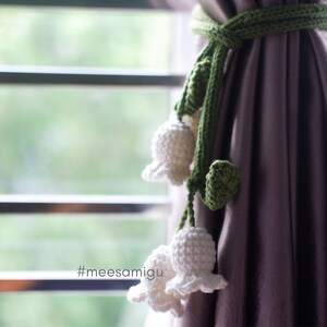 May include: A green crocheted curtain tie with white crocheted lily of the valley flowers. The tie is attached to a purple curtain. #meesamigu