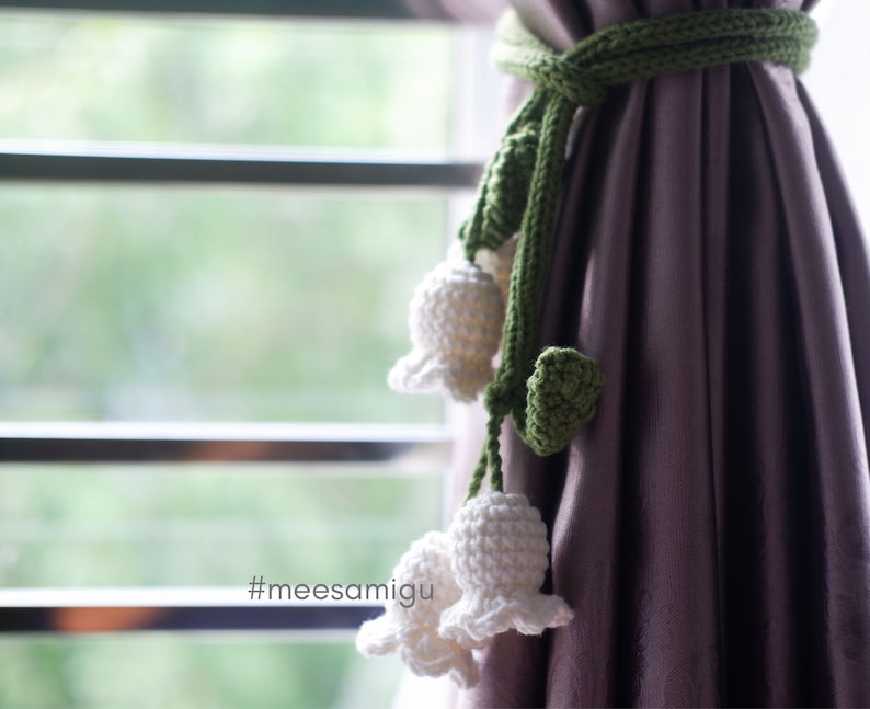 May include: A green crocheted curtain tie with white crocheted lily of the valley flowers. The tie is attached to a purple curtain. #meesamigu