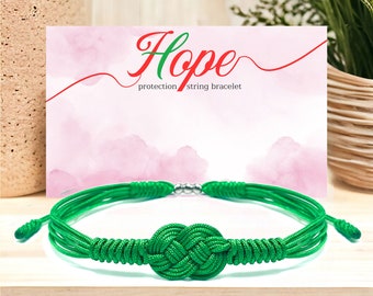 May include: Green string bracelet with a knot in the center. The bracelet is on a wooden surface. The text "Hope protection string bracelet" is on a pink card behind the bracelet.