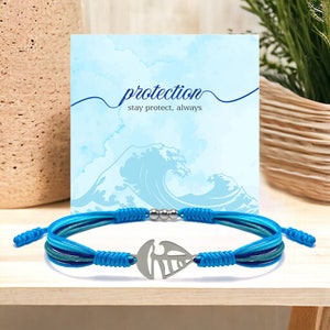 Peut inclure: Un bracelet en cordon tressé bleu et vert avec un pendentif en forme de feuille argentée. Le bracelet est doté d'une perle argentée à chaque extrémité. Le bracelet est posé sur une surface en bois avec une carte qui dit "protection stay protect, always" et une image aquarelle d'une vague.