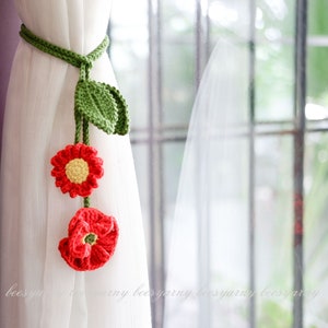 以下が含まれることがあります： 赤い花と緑の葉が付いた、かぎ針編みのカーテン留め。白いカーテンにかけられています。
