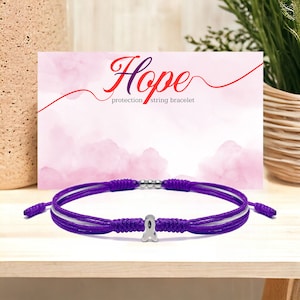 May include: Purple string bracelet with a silver ribbon charm. The bracelet is on a white surface with a pink background. The text "Hope protection string bracelet" is on the background.
