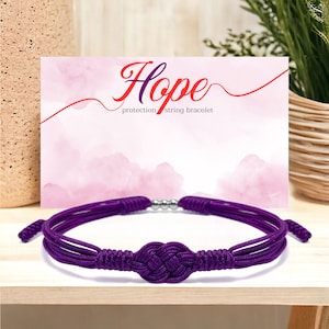 May include: Purple string bracelet with a knot in the center. The bracelet has a silver bead on each side of the knot. The bracelet is on a wooden surface. The card behind the bracelet says "Hope protection string bracelet".