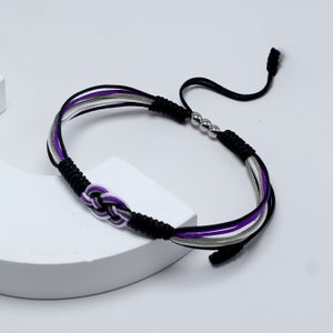 Op de afbeelding: Een gevlochten armband in zwart, paars, wit en grijs. De armband heeft een geweven knoopontwerp in het midden en verstelbare schuifknopen. Het heeft drie zilveren kralen en ligt op een wit oppervlak.