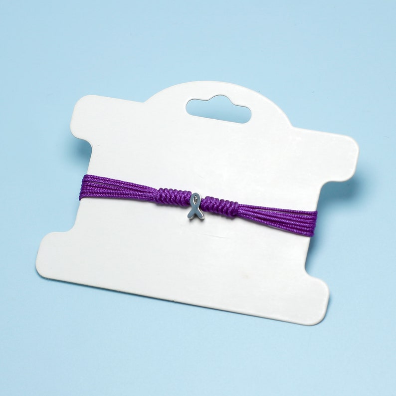 K&ouml;nnte beinhalten: Lilafarbenes Armband aus Schnur mit einem silbernen Band-Anh&auml;nger auf einer wei&szlig;en Karte.