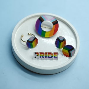 Può includere: Una collezione di gioielli a tema Pride, tra cui una spilla a righe arcobaleno, un paio di orecchini a bottone, un anello e la parola "PRIDE" in acrilico trasparente. I gioielli sono esposti su un vassoio rotondo bianco.