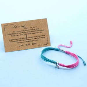 May include: A teal and pink string bracelet with a silver charm in the shape of a ribbon. A small card with the text "Life is tough but so are you!" is next to the bracelet. The card also includes the words "LOVE - HOPE - LIFE".