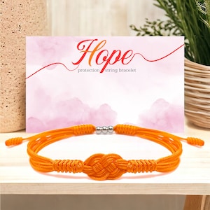 Może przedstawiać: Pomarańczowa bransoletka ze sznurka z węzłem w środku. Bransoletka leży na drewnianej powierzchni. Karta za bransoletką głosi "Hope protection string bracelet".