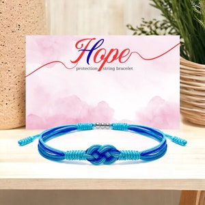 Puede incluir: Pulsera de cuerda azul y turquesa con una cuenta plateada y un nudo en el centro. La pulsera está sobre una superficie de madera con un fondo rosa y blanco que dice "Hope protection string bracelet".