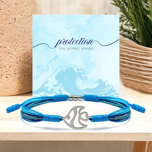 Op de afbeelding: Blauw en groen verstelbaar koordarmband met een zilveren visbedel. De armband heeft een kaartje eraan bevestigd met de tekst "protection stay protect, always" met een aquarel golfontwerp op de achtergrond.