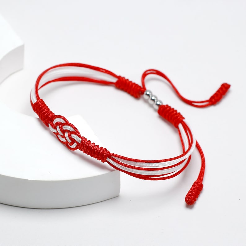 May include: Red and white braided bracelet with a silver bead accent. The bracelet is tied with a knot.