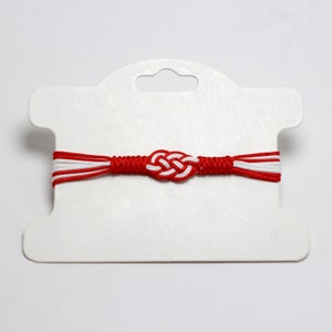 May include: Red and white braided bracelet with a knotted design. The bracelet is displayed on a white card with a hole for hanging.