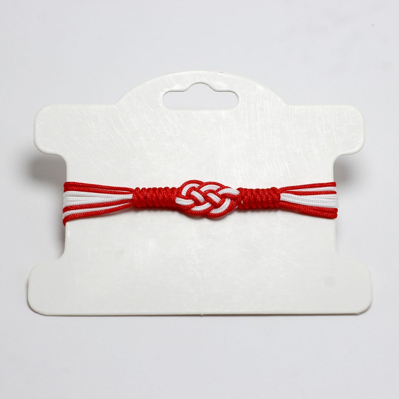 May include: Red and white braided bracelet with a knotted design. The bracelet is displayed on a white card with a hole for hanging.