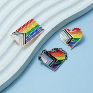May include: Three enamel pins featuring the Progress Pride flag. The first pin is a rectangular flag, the second is a heart-shaped flag, and the third is a heart-shaped flag with the text "You can be yourself with me".
