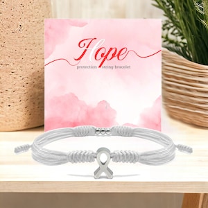 May include: White string bracelet with a silver ribbon charm. The bracelet is on a white surface with a pink background. The text "Hope protection string bracelet" is on the pink background.