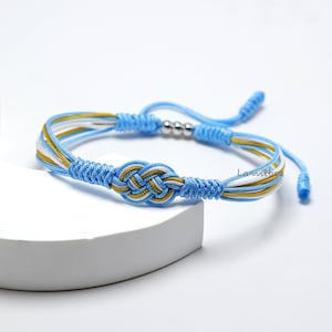 May include: A blue, white, and gold braided friendship bracelet with a knot in the center. The bracelet has silver beads and adjustable closures.