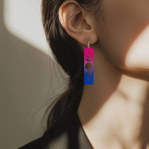 May include: A pair of dangle earrings with a pink to blue ombre gradient and a moon phase design. The earrings are made of metal and have a silver hook.