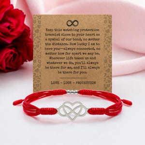 May include: A red string bracelet with a silver infinity heart charm. The bracelet is displayed with a card that has text about love, luck, and protection. A red rose is in the background.