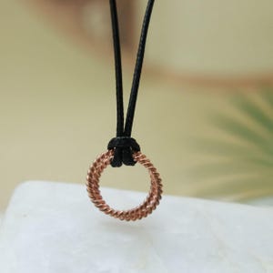 Op de afbeelding: Een cirkelvormige hanger gemaakt van gedraaid koperkleurig draad, hangend aan een dun zwart koord. De metalen ring heeft een getextureerd, gevlochten uiterlijk en is met een eenvoudige lusknoop aan het koord bevestigd voor een strak ontwerp.