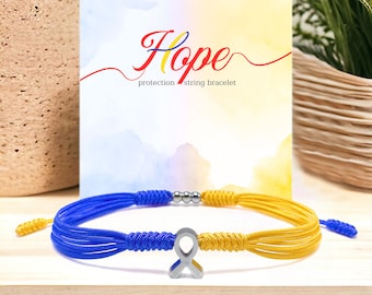 May include: A blue and yellow string bracelet with a silver ribbon charm. The bracelet is displayed in front of a card with the word "Hope" and the words "protection string bracelet". The bracelet is on a wooden surface.