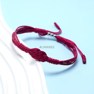 May include: A red braided bracelet with silver beads and a knot in the center. The bracelet has a tassel on each end. #LAMIBEE