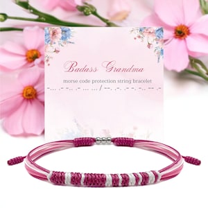 Op de afbeelding: Een roze en witte touw armband met een zilveren sluiting wordt getoond voor een kaart met de tekst "Badass Grandma" met de woorden "morse code protection string bracelet" eronder. De achtergrond toont roze bloemen.