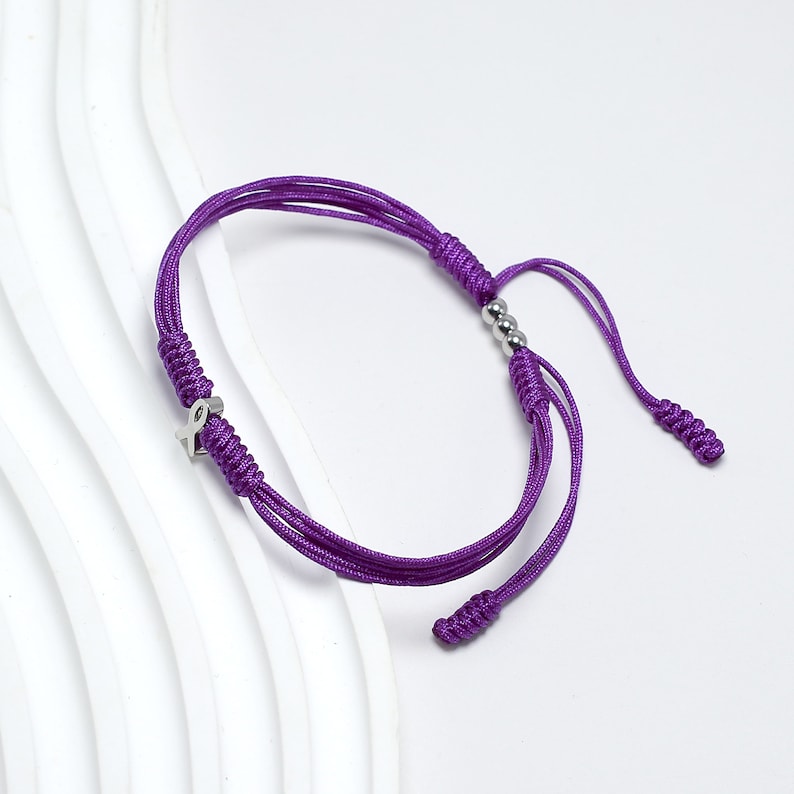 K&ouml;nnte beinhalten: Lilafarbenes geflochtenes Armband mit einem silbernen Schleifenanh&auml;nger und silbernen Perlen.