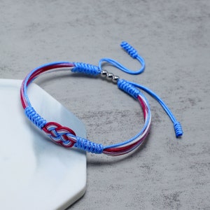 Puede incluir: Un brazalete de amistad trenzado azul, blanco y rojo con un acento de cuentas plateadas. El brazalete tiene un diseño de nudo y está atado con un nudo simple.