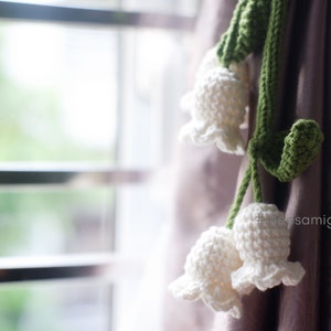 May include: A white crocheted lily of the valley flower garland with green leaves hanging from a curtain.