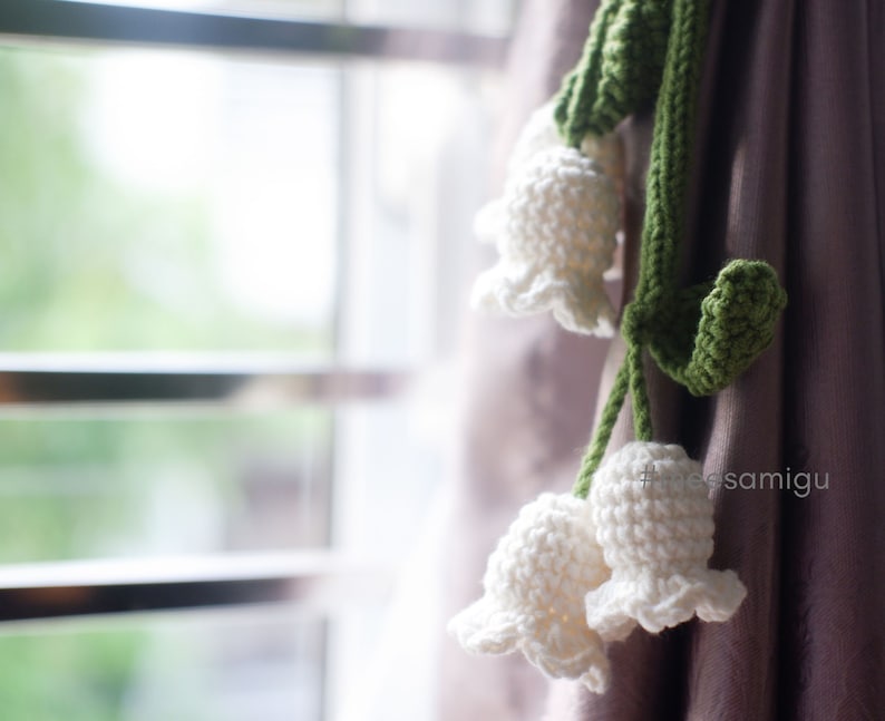 May include: A white crocheted lily of the valley flower garland with green leaves hanging from a curtain.
