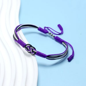 Könnte beinhalten: Ein geflochtenes Armband in Lila und Weiß mit einem silbernen Perlenakzent. Das Armband hat ein Knotendesign und ist mit einem Quastenverschluss versehen.