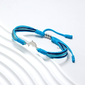 Puede incluir: Un brazalete de cordón trenzado azul y turquesa con un dije de delfín plateado y cuentas plateadas. El brazalete es ajustable con un nudo deslizante.