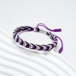 May include: A purple, black, and white braided bracelet with a silver bead accent. The bracelet has a knotted tassel on each end.