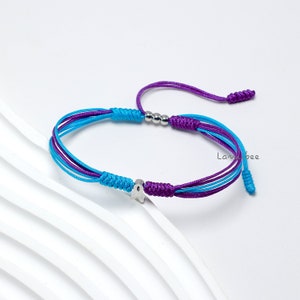 Puede incluir: Pulsera hecha a mano con un dije de estrella y cierre de nudo corredizo. La pulsera está hecha de cordones azules, morados y blancos. El dije de estrella es de color plateado. La pulsera se muestra sobre una superficie blanca.