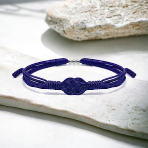 Puede incluir: Una pulsera trenzada de color morado oscuro con un diseño anudado. La pulsera está hecha de varios hilos de cordón y tiene un acento de cuentas plateadas.